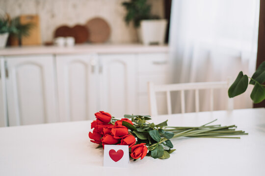 Bouquet of red roses and small card with painted red heart on white table.