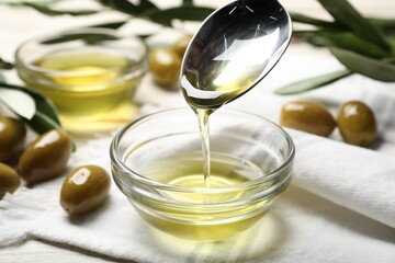 Spoon with cooking oil over bowl and olives on white wooden table, closeup