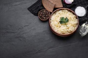 Bowl of tasty sauerkraut and ingredients on black table, flat lay. Space for text