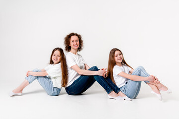 Mom and two teenager daughters in jeans and white t-shirts are smiling and happy. They sit with their knees bent. White background.