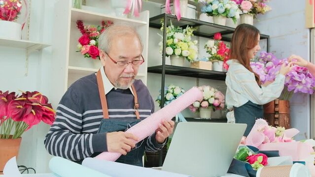 Asian elderly male florist owner online lives streaming for selling in e-commerce business website, employee and customer with blossom behind, working in colorful flower shop store, SME entrepreneur.
