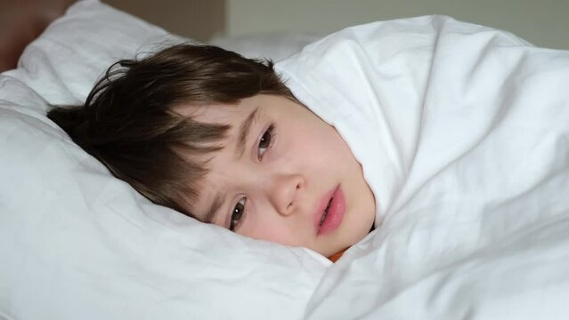 One unhappy boy in bed under blanket. The child does not want to fall asleep during the daytime. White linens. A person feeling bad. The baby is ill. After a bad dream. An orphan. Loneliness concept.