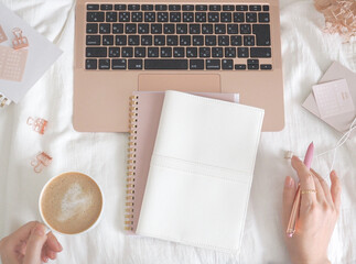 ベッドの上にラップトップとノート、カフェラテを持つ女性の手、柔らかな雰囲気のPCイメージ