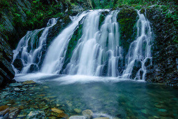 Fototapeta na wymiar Beautiful mountain rainforest waterfall with fast flowing water and rocks, long exposure. Natural seasonal travel outdoor background with sun shining. Stream waterfall on rocks in the forest