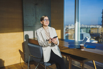 Cute positive female employee with long blond hair in casual clothes looking at phone while working on new business project at table with laptop and gadgets in office