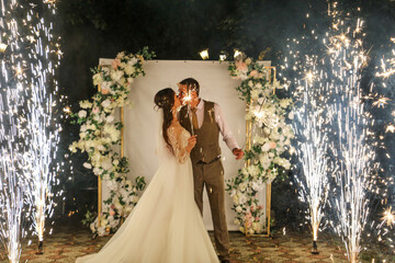 The bride and groom in a wedding dress in the sparks of fireworks at night.