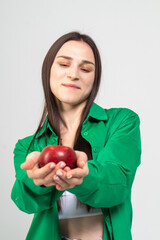 A cute brown-haired girl holds a red ripe apple in her hands on a white background. The model is wearing a green shirt and blue jeans.