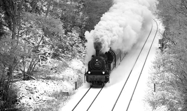 Steam train on the “Ruhrtalbahn“ (Ruhr valley line) between Arnsberg and Meschede Sauerland Germany. Snowy winter landscape panorama with big puff of smoke from historic locomotive.