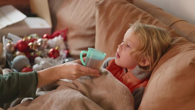 Close up, sick child gets warm drink while lying on the sofa with fever, wipes her mouth with blanket, mothers helping hand, caring moment.Corona symptoms, virus disease, ilness concept.HQ 4K footage