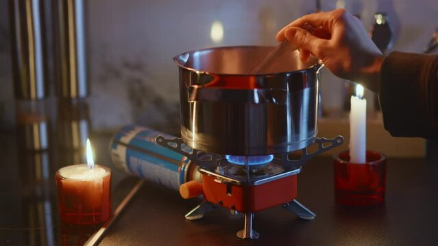 Portable stove usage at home during blackout, cooking on a tourist gas burner, stiring dish in a steel pot. Beauiful domestic background, burning candles. Surviving in Ukraine during war. HQ 4k