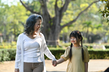Smiling grandmother and her little grandchild walking together surrounded by trees during sunny day