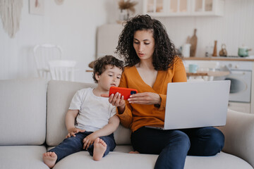 Young curly Italian Woman in orange blouse sits with son on couch shows him how to use mobile phone for gaming, remote works uses laptop. Home office. Cute little boy watching cartoons on cell phone.