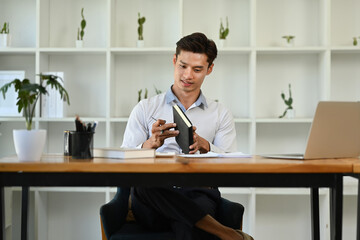 Fototapeta premium Smiling millennial businessman sitting at his desk in a bright modern office and using laptop