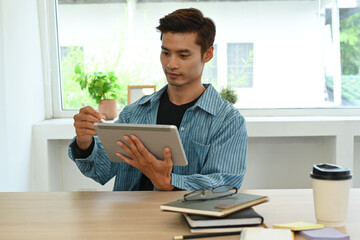 Handsome asian male freelancer checking online information on digital tablet, working on modern home office