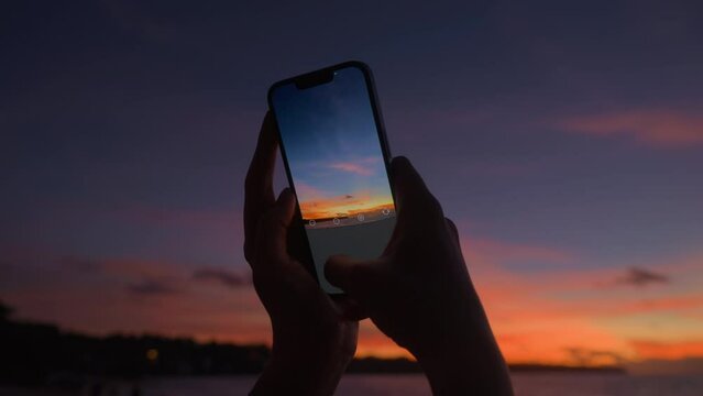 A girl photographs a colorful tropical sunset on a modern close-up phone. Silhouette of a woman's hands taking a photo of a beautiful orange sunset sky. Film grain texture.