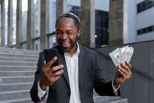 African American Man In Business Suit Rejoices, Got Online Small Business Loan, Owner With Money Cash Dollars Happy With Achievement Result, Boss Holding Cash And Phone.