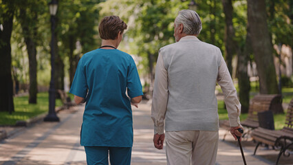 Elderly man with walking stick talking to medical employee in hospital park, back view