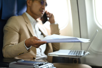 Businessman holding financial report and talking on mobile phone while sitting near windows on airplane during flight