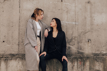 Two pretty women friends posing near gray wall. Couple of gay lesbian girls hugging embracing together girlfriends, dressed casual outfits, have a date. LGBT concept. Fashion, make up, hairstyle

