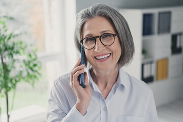 Photo of beautiful positive aged lady banker speak communicate telephone coworking workplace indoors