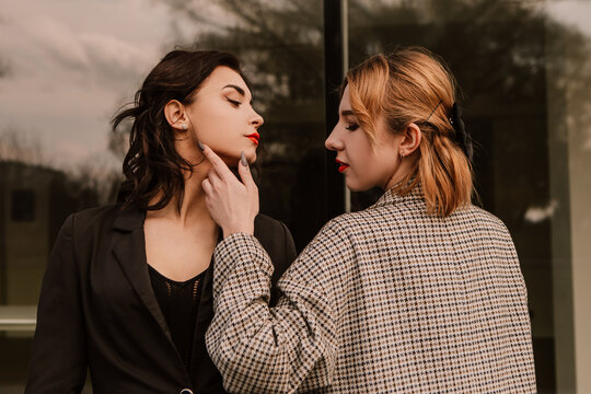 Two pretty women friends posing near glass building. Couple of gay lesbian girls hugging embracing kissing girlfriends, dressed casual outfits, have a date. LGBT concept. Fashion, make up, hairstyle
