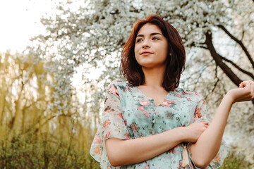 Portrait of charming pretty woman dressed flowery dress posing near apple cherry tree blossoms blooming flowers in the garden park in early spring nature. Fashion, girl model with black hair
