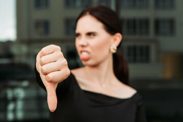 Pretty brunette caucasian woman standing outside street with white teeth beaming healthy lady. Happy millennial girl showing thumb down dislike disapprove sign gesturing making faces show tongue
