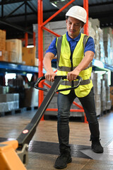 Young man storehouse worker in safety hardhats pulling a pallet truck through rows of storage racks with merchandise