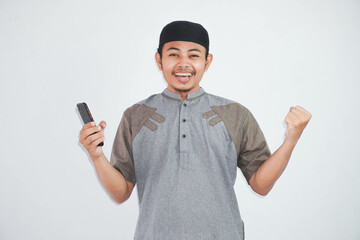 Excited or happy young Asian Muslim man holding on mobile phone and hands in fists doing winner gesture wearing koko clothes isolated over white background