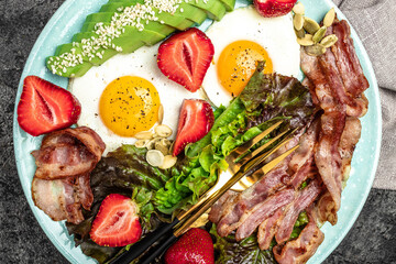 Plate with fried eggs, bacon, avocado, strawberries and fresh salad on a dark background. Ketogenic diet food, healthy meal concept, top view