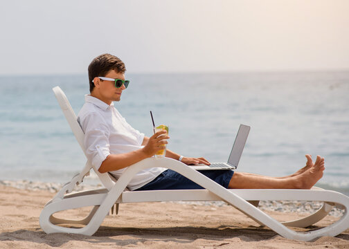 Young man in sunglasses and white shirt is lying on a beach chair on the seaside holding a cocktail and a laptop.