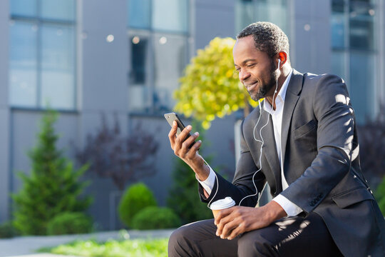 African American Businessman Using Phone Sitting On Bench Outside Office Building, Boss Watching Video And Smiling In Headphones, Man Outdoors Listening Music Audiobook And Podcasts Online Radio.