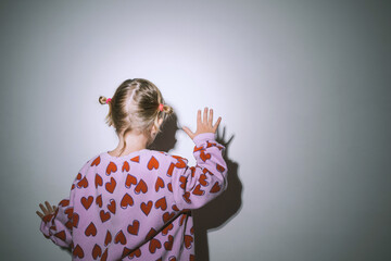 Photo of little girl dancing and playing with her shadow against bright light against white wall.