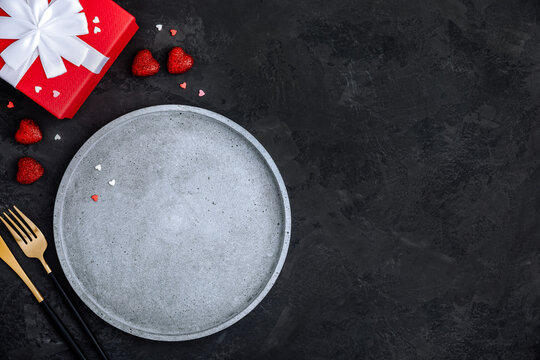 Valentine's Day table setting with empty plate, red hearts and present box on dark background