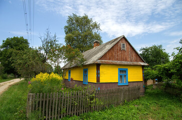 Fototapeta na wymiar A bright rural wooden house of yellow color behind a fence among flowers against a blue sky.