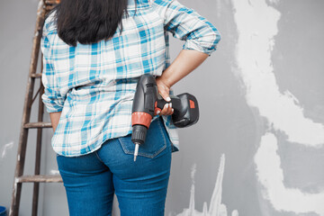 Cropped shot of back of a woman holding drill on her hips while looking at wall