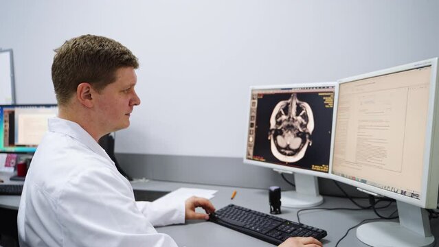 Male technician in hospital lab sitting in front of computer. Attentive medic looks at head scan analyzing the image and diagnosing.
