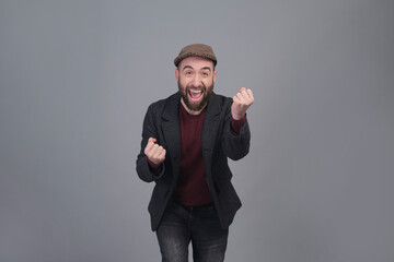 Enthusiastic young man in jacket and cap with arms up in a studio shot - 565303085