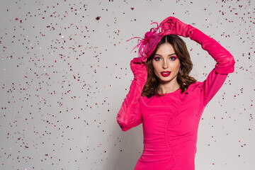 young brunette woman in magenta color dress adjusting elegant hat on grey with falling confetti.