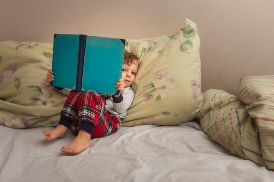Toddler in pajamas sitting on bed and reading bedtime book looki