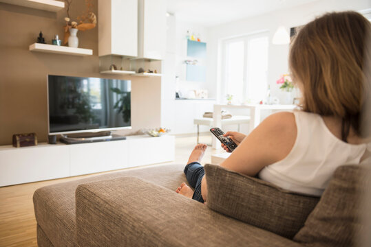 Pregnant woman sitting on sofa and watching TV with remote control, Munich, Bavaria, Germany