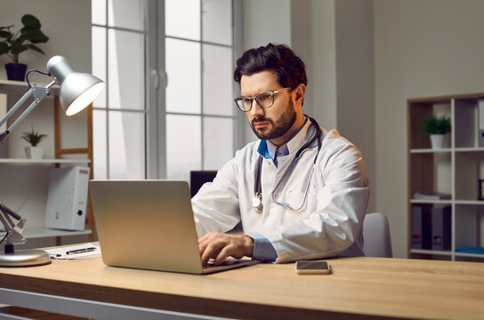 Professional male doctor filling documents using laptop computer. Medical expert wearing glasses and white lab coat taking notes in medical journal, checking electronic files of patients
