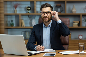 Portrait of successful smiling man in office, mature businessman looking at camera cheerfully, senior boss in glasses and beard working inside office with laptop and documents, signing contract.
