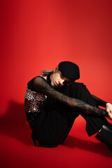 tattooed queer model in black elegant attire sitting and looking at camera on red background.