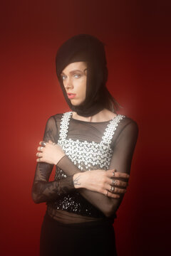 nonbinary person in black headwear and top with sequins posing with crossed arms on dark red background.