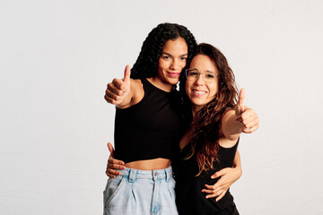 Two close friends showing their thumbs up to the camera while smiling at a studio shot - 565320644