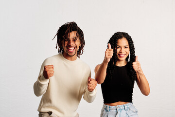 A young latin couple expressing happiness with thumbs up and clenched fists smiling at the camera in a studio shot - 565320659