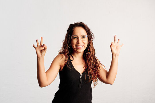 An adult woman making the ok gesture with both hands and looking at the camera in a studio