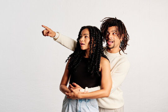 A young man hugging his girlfriend from behind as he points to a copy space with amazement expression in a studio shot