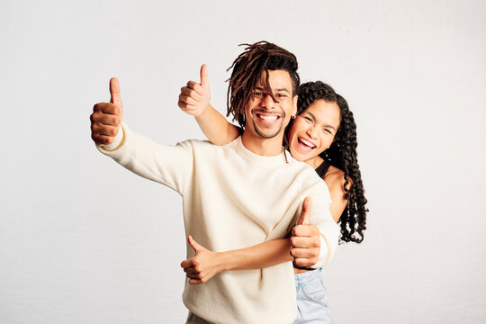 A cheerful young woman behind her cheerful boyfriend making two thumbs-up with hands in a studio